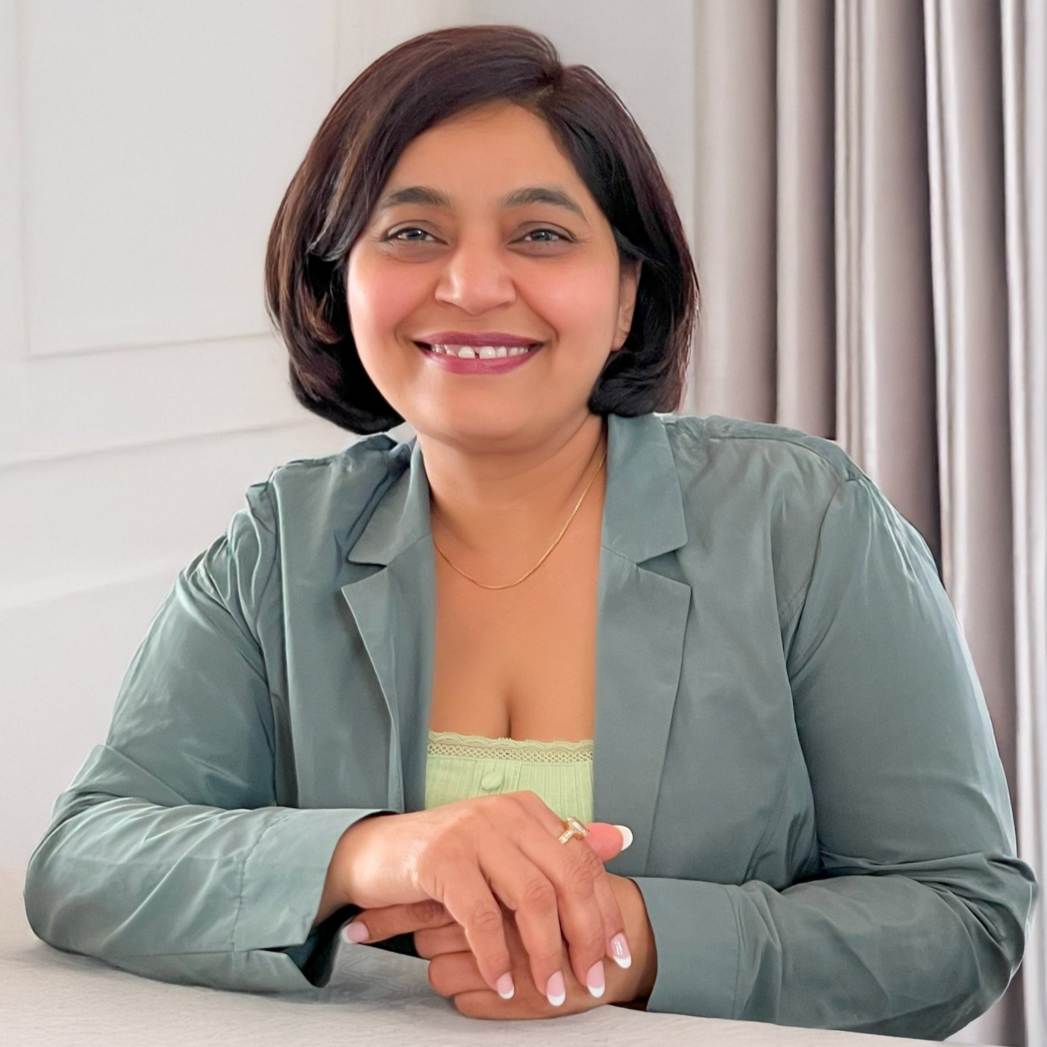 Portrait einer lächelnden Frau mit dunklen Haaren im grünen Blazer, sitzend an einem Tisch vor hellem Hintergrund.