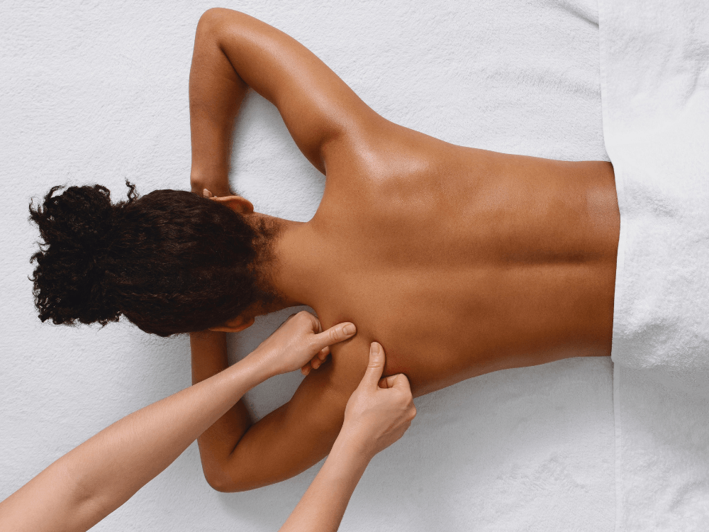 Frau liegt auf dem Bauch und erhält eine Rückenmassage, Therapeutin massiert mit beiden Daumen gezielt die Schultermuskulatur.