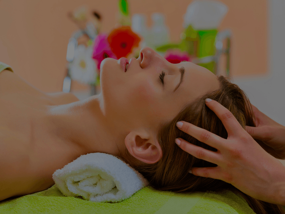 Frau erhält eine ayurvedische Kopfmassage im Liegen, entspannte Atmosphäre mit Blumen im Hintergrund.